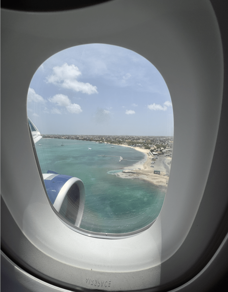 View from an airplane window showing a turquoise sea, sandy beach, and coastal buildings under a partly cloudy sky, with the airplane’s engine visible on the left—perfect inspiration for your next travel blog or meaningful travel guide.